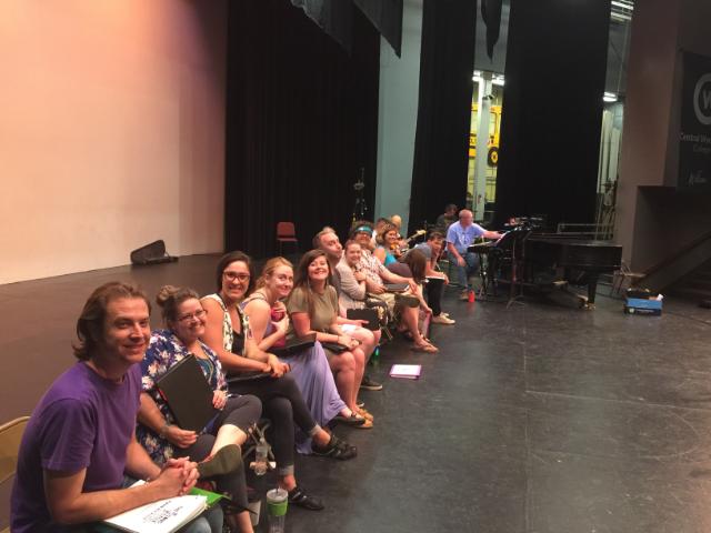 The cast of the retail song cycle sits on stage ready for rehearsal. 